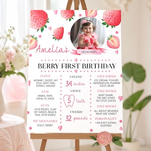 Op de afbeelding: Een wit verjaardagsbord met roze aardbeienillustraties, een ronde kinderfoto en tekst waaronder Amelia's Berry First Birthday, 34 inches, 5 teeth en 32 pounds, weergegeven op een houten ezel met roze bloemaccenten.