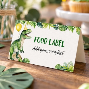 Puede incluir: Una tarjeta blanca plegada con un dinosaurio verde que lleva un gorro de fiesta amarillo y sostiene un globo amarillo. La tarjeta presenta ilustraciones de hojas tropicales verdes, el texto FOOD LABEL en verde y Add your own text en negro.