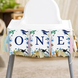 May include: A high chair banner featuring three white pennants with the dark blue letters O, N, and E. Each pennant displays watercolor illustrations of sea creatures including whales, sea turtles, and fish, accented with pastel fringe tassels.