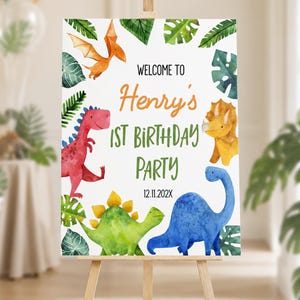 Op de afbeelding: Een wit rechthoekig verjaardagsbord met aquarelillustraties van een rode, gele, groene en blauwe dinosaurus en een oranje pterosauri&euml;r. De tekst luidt: WELCOME TO Henry's 1ST BIRTHDAY PARTY 12.11.202X, omringd door groene tropische bladeren.