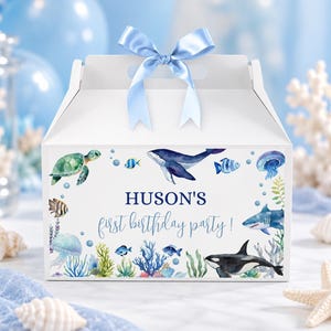 May include: A white cardboard gable box with a light blue satin ribbon bow on top. The front features watercolor illustrations of sea creatures including a turtle, whale, shark, orca, and jellyfish, with the text HUSON'S first birthday party!