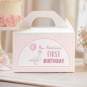 May include: A white gable box featuring a light pink gingham pattern and a central arch graphic. The design includes a watercolor illustration of a goose wearing a party hat and holding a pink balloon with the number 1, and the text Amelia's FIRST BIRTHDAY.