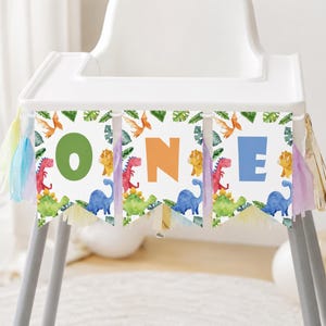 K&ouml;nnte beinhalten: Ein Hochstuhl-Banner bestehend aus drei wei&szlig;en Wimpeln mit Aquarell-Illustrationen von Dinosauriern und tropischen Bl&auml;ttern. Die Buchstaben O, N und E sind in gro&szlig;en gr&uuml;nen, orangen und blauen Schriftarten gedruckt. Mit bunten Quasten.