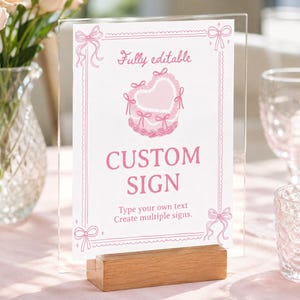 Peut inclure: Un porte-affiche en acrylique transparent avec une base en bois pr&eacute;sentant une carte blanche avec du texte et des graphismes roses. La carte comporte une illustration de g&acirc;teau en forme de c&oelig;ur, des n&oelig;uds roses et le texte Fully editable, CUSTOM SIGN.