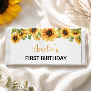 May include: A rectangular candy bar wrapper featuring a watercolor sunflower border along the top edge. The white label displays the text Amelia's in a gold script font and the words FIRST BIRTHDAY in a bold black sans-serif typeface.