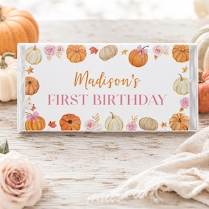 May include: A rectangular white sign featuring the orange text Madison's and the pink text FIRST BIRTHDAY. The border is decorated with watercolor illustrations of orange and white pumpkins, pink roses, and small autumn leaves in shades of red and gold.