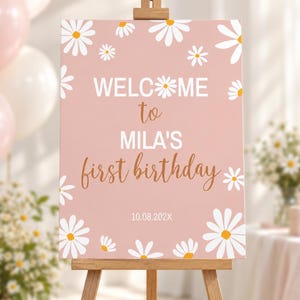 May include: A rectangular pink sign on a wooden easel featuring white daisy illustrations with yellow centres. The text reads WELCOME to MILA'S first birthday 10.08.202X. The design includes white daisies scattered around the border of the sign.