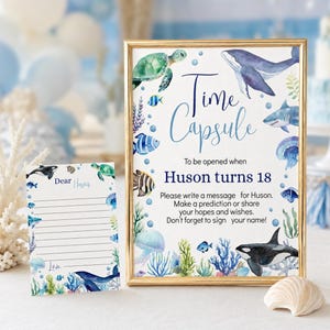 Pu&ograve; includere: Un cartello con cornice dorata e un biglietto abbinato con illustrazioni ad acquerello di vita marina, tra cui una balena, una tartaruga, uno squalo e una medusa. Il cartello recita Time Capsule To be opened when Huson turns 18. Il biglietto dice Dear Huson.