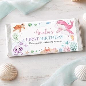 May include: A rectangular candy bar wrapper featuring watercolor illustrations of a sea turtle, jellyfish, octopus, crab, seahorse, and pink whale. The text reads Amelia's FIRST BIRTHDAY Thank you for celebrating with me! in pink and blue script.