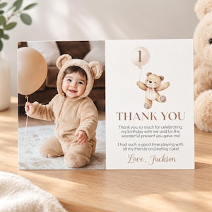 May include: A rectangular thank you card featuring a photo of a baby in a tan bear-ear jumpsuit holding a beige balloon. The right side displays a watercolor illustration of a teddy bear with a balloon, the number 1, and the text Thank You.