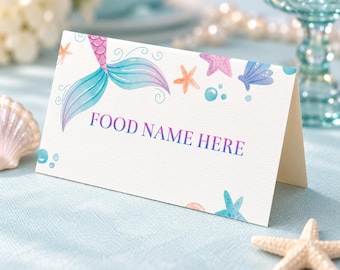 May include: A white folded card with a watercolor mermaid tail, starfish, seashells, and bubbles printed on the front. The text FOOD NAME HERE is printed in purple in the center. The card has a textured surface and a clean, simple design.