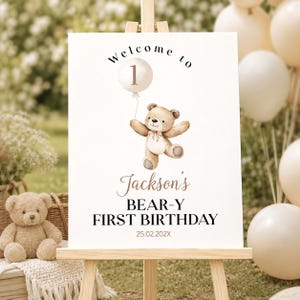 May include: A white rectangular sign on a wooden easel featuring a watercolor illustration of a brown teddy bear holding a balloon with the number 1. The text reads Welcome to Jackson's BEAR-Y FIRST BIRTHDAY and the date 25.02.202X in black.