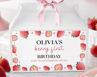 May include: A white gable box with a pink satin ribbon bow on top. The front features a strawberry border and the text: OLIVIA'S berry first BIRTHDAY THANK YOU FOR CELEBRATING WITH ME! The design includes watercolor-style strawberry illustrations.