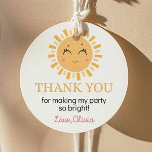 May include: A round white gift tag featuring a yellow sun illustration with a smiling face and pink cheeks. The text reads THANK YOU for making my party so bright! Love, Olivia. The tag is attached with white string to a brown paper wrapped package.