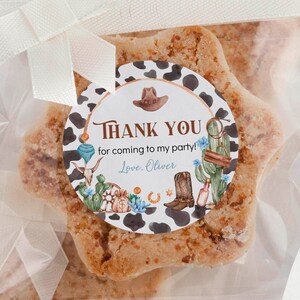 May include: Star-shaped cookies in clear plastic bags tied with white ribbons. Each bag features a round sticker with a cow print border, a brown cowboy hat, a boot, cacti, and the text: THANK YOU for coming to my party! Love, Oliver.