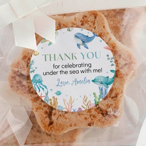 Op de afbeelding: Een transparante cellofaan zak met een wit satijnen strik, die een stervormige suikerkoek bevat die is versierd met een ronde sticker. De sticker heeft een aquarel-illustratie van zeeleven, waaronder een blauwe walvis, een kwal en koraal. De tekst op de sticker luidt "Bedankt voor het vieren onder de zee met me! Liefs, Amelia."