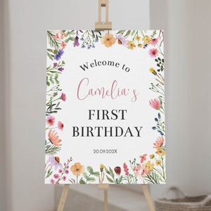 K&ouml;nnte beinhalten: Ein rechteckiges Schild auf einer Holzstaffelei mit einem Aquarell-Blumenrand aus rosa, gelben, orangen und violetten Wildblumen. Der Text lautet Welcome to Camelia's FIRST BIRTHDAY 20.09.202X in schwarzen und rosa Schriftarten.