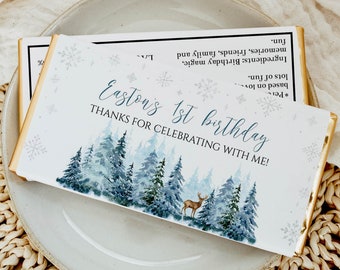 May include: A rectangular chocolate bar wrapper featuring a watercolor forest of blue and green pine trees, a small deer, and snowflakes. The text reads: Easton's 1st birthday, THANKS FOR CELEBRATING WITH ME! The back lists ingredients like birthday magic.