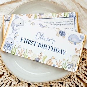 May include: A personalized chocolate bar with a marine theme, featuring illustrations of a dolphin, whale, and octopus. The text "Oliver's FIRST BIRTHDAY" is displayed in blue. The bar rests on a gray plate.