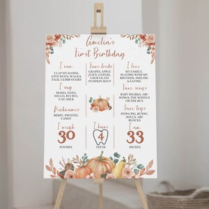 May include: A white birthday milestone sign featuring orange pumpkin illustrations, floral accents, and text detailing Amelia's first birthday facts including weight in pounds, height in inches, favorite foods, songs, toys, and nicknames.