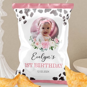 May include: A personalized chip bag with a pink and white cow print design. The bag features a photo of a baby girl, the name "Evelyn's", and the text "1st Birthday" with a date. Two potato chips are in the foreground.