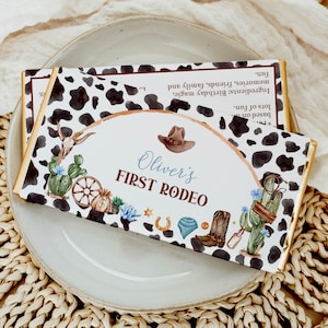 May include: A white and black cow print chocolate bar wrapper with gold trim. The wrapper has a brown, gold, and blue illustration of a cowboy hat, cactus, and horseshoe. The text on the wrapper reads "Oliver's First Rodeo".