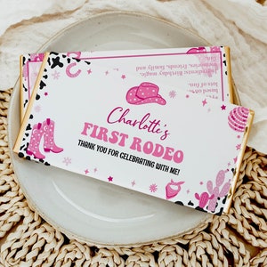 May include: Pink and white chocolate bar wrapper with a pink cowboy hat, boots, and cacti. The text reads "Charlotte's First Rodeo" and "Thank you for celebrating with me!"