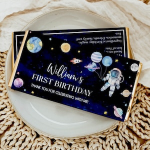 Puede incluir: Una barra de chocolate azul y negro con temática espacial. Presenta un astronauta, planetas, estrellas y el texto "William's First Birthday. Thank you for celebrating with me!"