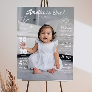 May include: A rectangular milestone birthday sign featuring a central photograph of a child in a white dress. The sign includes white text and graphics detailing milestones such as 28 inches, 30 pounds, 5 teeth, and the text Amelia is One.