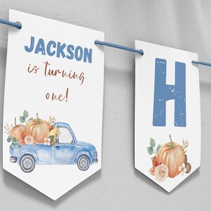 May include: A white decorative banner featuring blue text that reads JACKSON is turning one! and a blue truck illustration carrying orange pumpkins and flowers. The banner includes additional panels with large blue letters and pumpkin graphics.