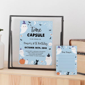 May include: A light blue time capsule sign and matching note card featuring Halloween illustrations of ghosts in hats, pumpkins, a black cat, and spiderwebs. The text reads Time Capsule to be opened on Mason's 18th birthday October 30th, 2040.