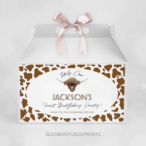May include: A white gable box with a pink satin ribbon on top. The front features a brown cow print border surrounding an oval with a Highland cow illustration and the text: Holy Cow, JACKSON'S First Birthday Party! THANK YOU FOR CELEBRATING WITH ME!