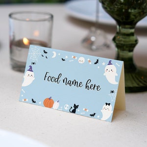 May include: A light blue folded card with the text Food name here in black script. The card features a Halloween-themed border with illustrations of white ghosts wearing purple hats, orange pumpkins, a black cat, spider webs, bats, and candy.