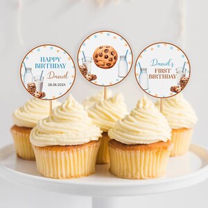 May include: Three round cupcake toppers featuring illustrations of milk bottles, blue striped straws, and chocolate chip cookies. The text reads HAPPY BIRTHDAY Daniel 28.08.202X and Daniel's FIRST BIRTHDAY. The toppers are inserted into frosted cupcakes.