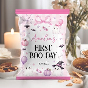 May include: A pink and white striped snack bag featuring the text Amelia's FIRST BOO-DAY and 18.10.202X. The design includes illustrations of ghosts, a witch hat, pumpkins, bats, a spider web, a bow, balloons, candies, and a bone.