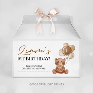 May include: A white gable box with a satin ribbon bow on top, featuring a printed label that reads Liam's 1ST BIRTHDAY! THANK YOU FOR CELEBRATING WITH ME! The design includes a brown teddy bear holding a cluster of balloons with the number 1.
