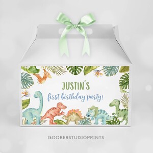 May include: A white gable box with a light green satin ribbon on top. The front features a watercolor illustration of various dinosaurs and tropical leaves surrounding the text "JUSTIN'S first birthday party!" in green and blue script.