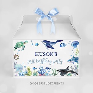 May include: A white gable box with a light blue satin ribbon on top, featuring a watercolor ocean theme with a sea turtle, whale, shark, orca, jellyfish, and various fish. The text reads HUSON'S first birthday party! in blue lettering.