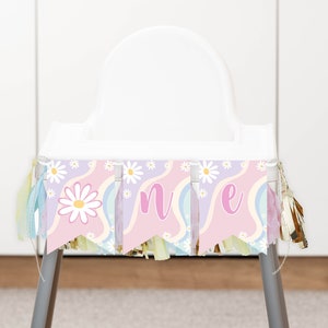 Op de afbeelding: Een kinderstoelbanner met drie wimpels op een golvende achtergrond in roze, lavendel en lichtblauw. De banner toont witte madeliefjes en de letters O, N en E in een roze schrijflettertype, geaccentueerd met hangende metallic gouden kwastjes.
