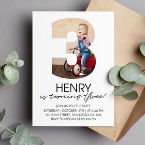 May include: A white birthday invitation card featuring a large number three filled with a photo of a child on a red toy car. The text reads: HENRY is turning three! JOIN US TO CELEBRATE SATURDAY | OCTOBER 15TH | AT 2:00 PM 123 MAIN STREET, SAN DIEGO, CA. 1345 RSVP TO MEGAN AT 123.456.789