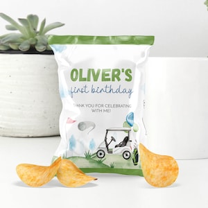 May include: A white packet of crisps with a green top and a golf theme. The packet says "Oliver's first birthday" and has a golf cart illustration. Three potato crisps are in front of the packet.