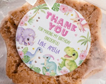 May include: A star-shaped cookie packaged in a clear plastic bag with a cream-colored ribbon. A circular sticker features illustrations of dinosaurs, palm leaves, and pink flowers with the text: THANK YOU FOR MAKING MY BIRTHDAY ONEDERFUL! LOVE, AMELIA.