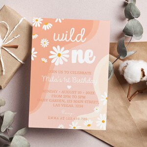 May include: A rectangular birthday invitation card featuring a peach and cream wavy background with white daisy illustrations. The text reads: wild one, JOIN US TO CELEBRATE Mila's 1st Birthday, SUNDAY AUGUST 10 202X FROM 2PM TO 5PM, DAISY GARDEN, 123 MAIN STREET, LAS VEGAS. 1234, RSVP EMMA AT 123.456.789.