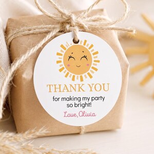 May include: A round white paper gift tag tied with twine to a brown paper wrapped box. The tag features a yellow sun illustration with a smiling face and the text: THANK YOU for making my party so bright! Love, Olivia, in orange and pink.