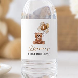 May include: A clear plastic water bottle with a white label featuring a brown teddy bear illustration holding a bunch of balloons with the number one inside one balloon. The text reads Liam's FIRST BIRTHDAY in brown and black script and print.