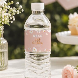 May include: A clear plastic water bottle with a white cap and a custom label. The label is light pink with white daisy illustrations and gold text that reads Mila's first birthday. The bottle is filled with clear water and has a ribbed texture.