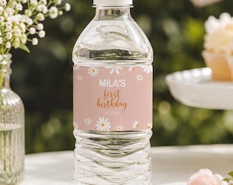 May include: A clear plastic water bottle with a white cap and a custom label. The label is light pink with white daisy illustrations and gold text that reads Mila's first birthday. The bottle is filled with clear water and has a ribbed texture.