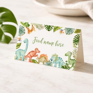 Może przedstawiać: Złożona biała kartka z akwarelową ilustracją r&oacute;żnych dinozaur&oacute;w i tropikalnych liści. Tekst "Food name here" jest nadrukowany zieloną kursywą na środku. Dinozaury obejmują niebieskie, pomarańczowe i zielone gatunki.
