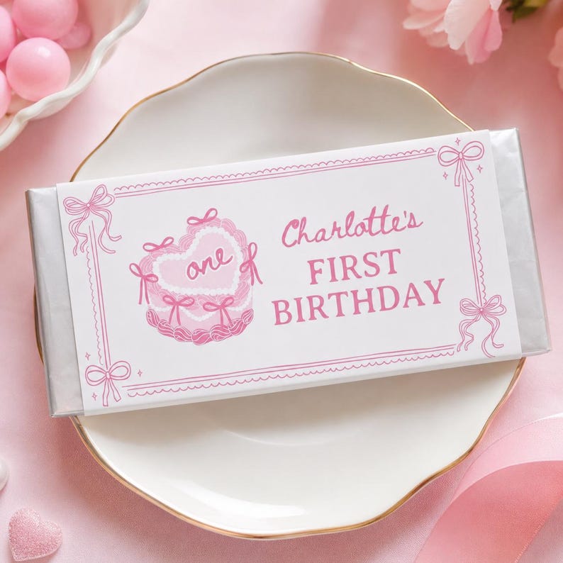 May include: A rectangular candy bar wrapper featuring a white background with pink line art. The design includes a heart-shaped cake with the word one, and the text Charlotte's FIRST BIRTHDAY, framed by decorative pink bows and borders.
