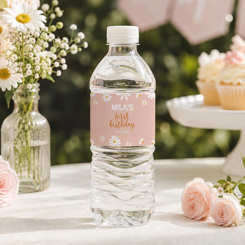 May include: A clear plastic water bottle with a white cap and a custom label. The label is light pink with white daisy illustrations and gold text that reads Mila's first birthday. The bottle is filled with clear water and has a ribbed texture.